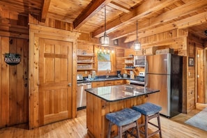 A wooden cabin interior with a kitchen featuring a central island, bar stools, a refrigerator, and a pantry sign.