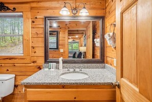 A wooden cabin bathroom with a granite countertop sink, a large mirror, and a window with blinds.