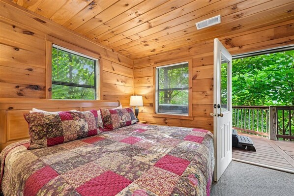 A wooden cabin bedroom with a large bed, two windows, a door leading to a deck, and a lamp on the nightstand.