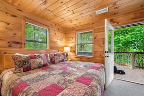 Un dormitorio de cabaña de madera con una cama grande, dos ventanas, una puerta que da a una terraza y una lámpara en la mesita de noche.