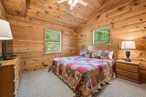 A wooden cabin bedroom with a bed, two windows, a ceiling fan, and a nightstand with a lamp.
