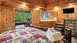 A wooden cabin interior with a bed, a desk, a chair, a television, and a view of greenery outside. - Tranquil Views w/ Mountain Views & Hot Tub (Sevierville)