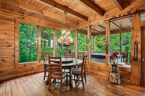A wooden cabin interior with a dining table, chairs, a chandelier, and large windows offering a view of the forest.