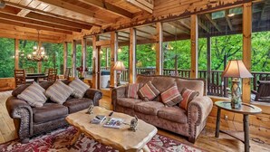 A wooden cabin with a large deck, furnished with leather sofas and a coffee table, and large windows offering a view of the forest. - Tranquil Views w/ Mountain Views & Hot Tub (Sevierville)
