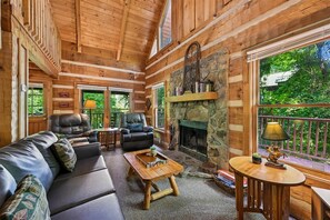 A log cabin interior with a fireplace, leather seating, and a balcony with a view of greenery.
