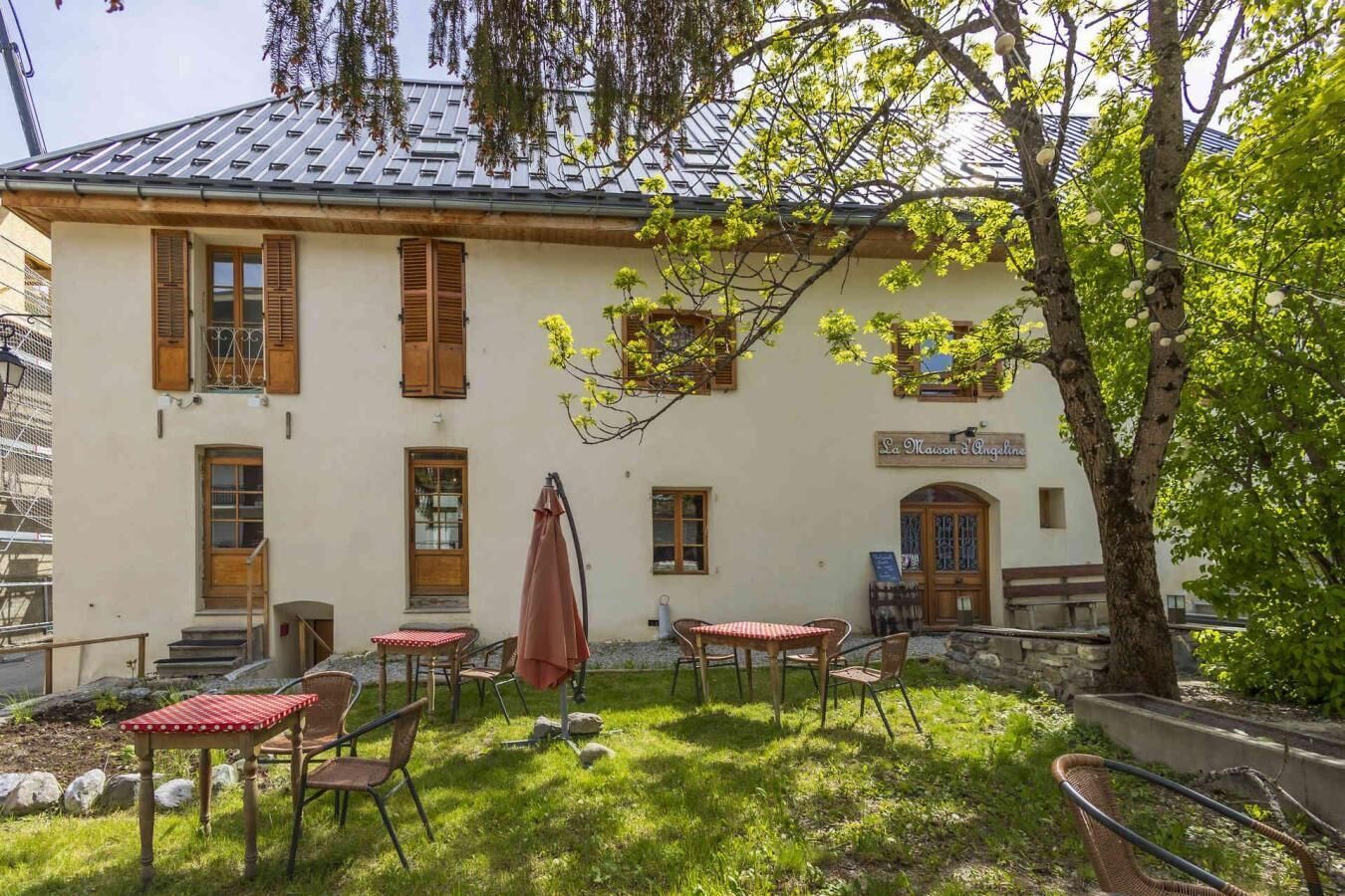 A building with a sign reading 'La Maison d'Angel'. There are outdoor tables and chairs, a tree, and a stone wall.