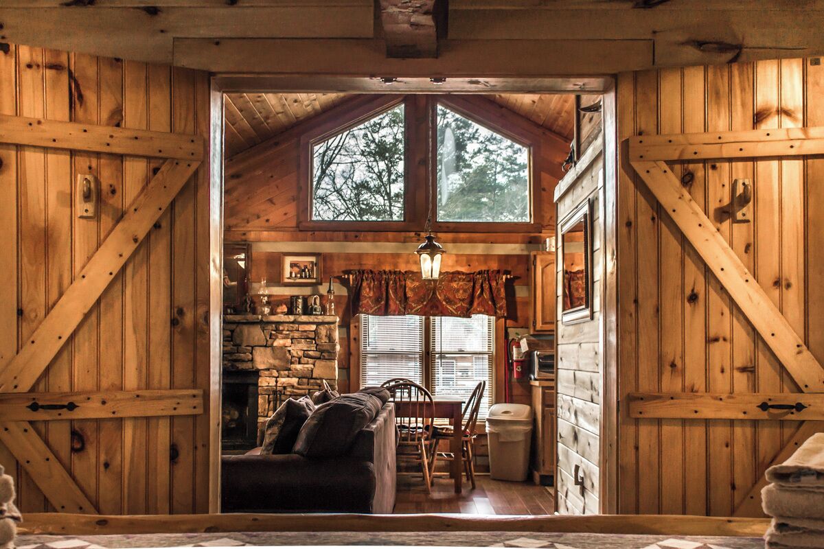 Interior rústico de una cabaña con chimenea de piedra, muebles de madera y ventanas amplias.