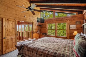 Ein Schlafzimmer in einer Holzhütte mit einem Bett, einem Deckenventilator, einem Fernseher und großen Fenstern.