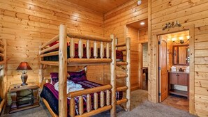 Una habitación tipo cabaña de madera con una cama litera, una mesita con una lámpara y una puerta que da a un baño.
