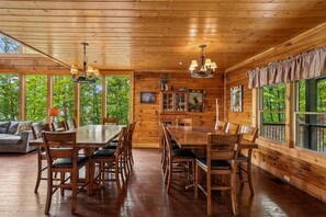 A wooden interior with a dining table, chairs, and a sofa. There are windows with curtains, a chandelier, and a cabinet with glass doors. - Amazing Views w/ Theater & Game Room (Sevierville)