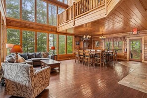A spacious wooden interior with a dining area, a sofa set, and large windows offering a view of the outdoors.
