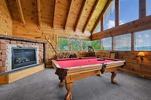 A wooden interior with a pool table, fireplace, and large windows.