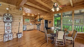 A wooden cabin interior with a dining area, kitchen, and large windows.