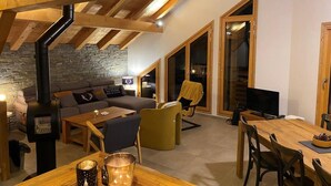 Una sala de estar acogedora con pared de piedra, vigas de madera, chimenea, sofá y un comedor con sillas y mesa.