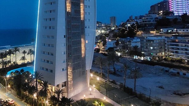 Ein hohes, modernes Hotel mit geschwungener Fassade in Strandnähe, mit Palmen und einer Straße.