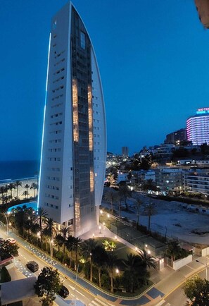 Um hotel alto e moderno com fachada curva, situado perto de uma praia com palmeiras e uma estrada.