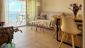 Ein modernes Wohnzimmer mit einer Couch, einem Esstisch und Stühlen. Ein Balkon bietet Blick auf das Meer.