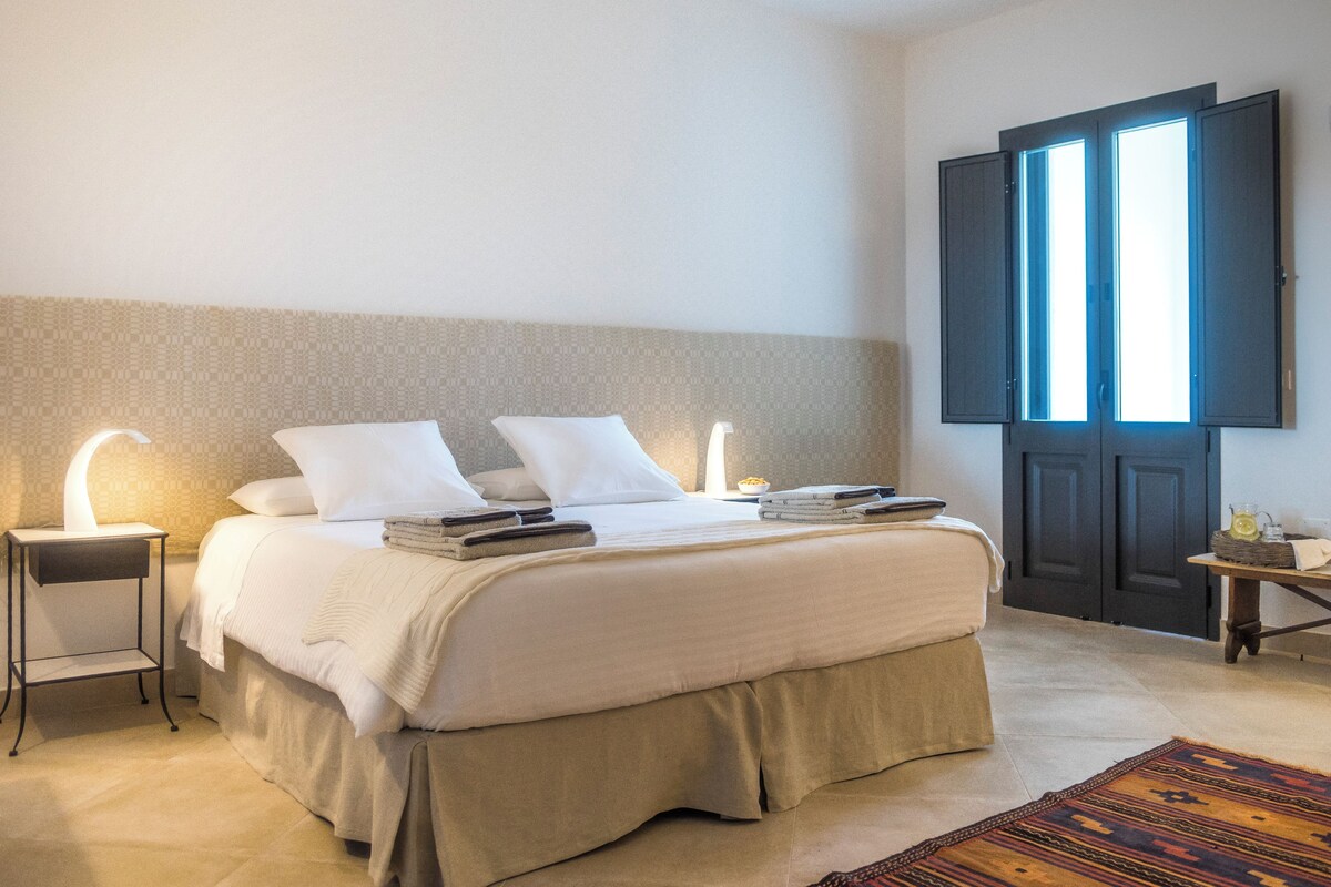 A neatly made bed with white linens, two bedside tables with lamps, a dark door, and a patterned rug on the floor.
