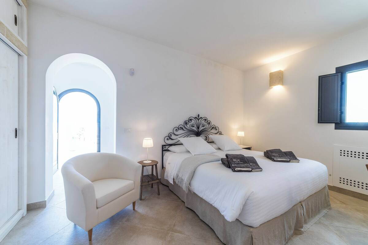 A bedroom with a bed, a chair, a small table, and a wall-mounted TV.