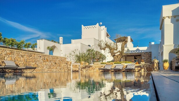 A pool area with lounge chairs, a stone wall, and a building with a white facade and arched windows.