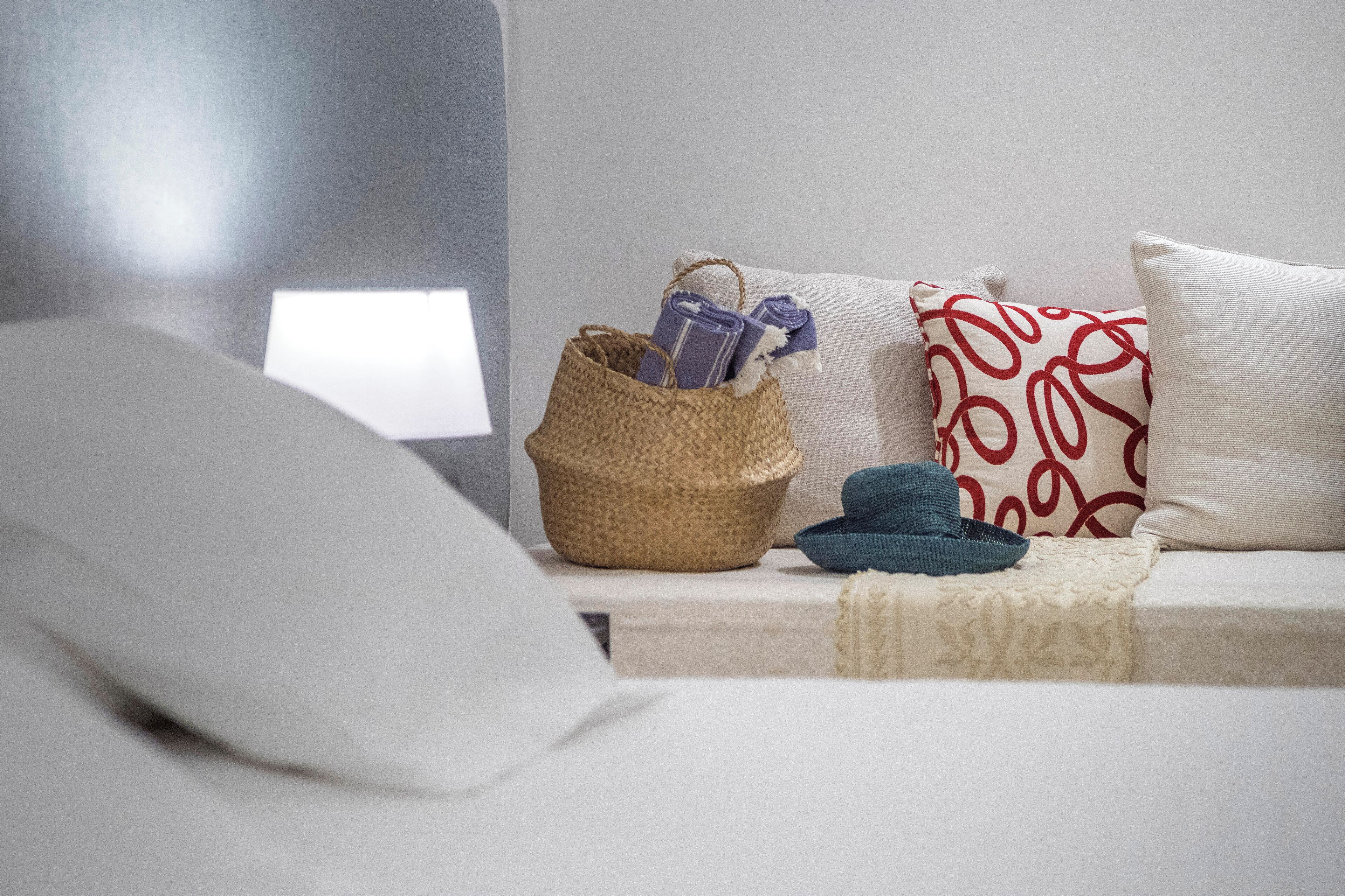 A neatly arranged bedroom with a bed, a bedside table with a lamp, and a woven basket containing folded towels and a hat.