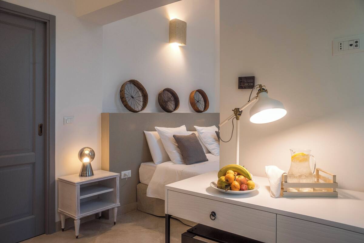 A neatly arranged bedroom with a bed, bedside table, lamp, and wall-mounted clocks.