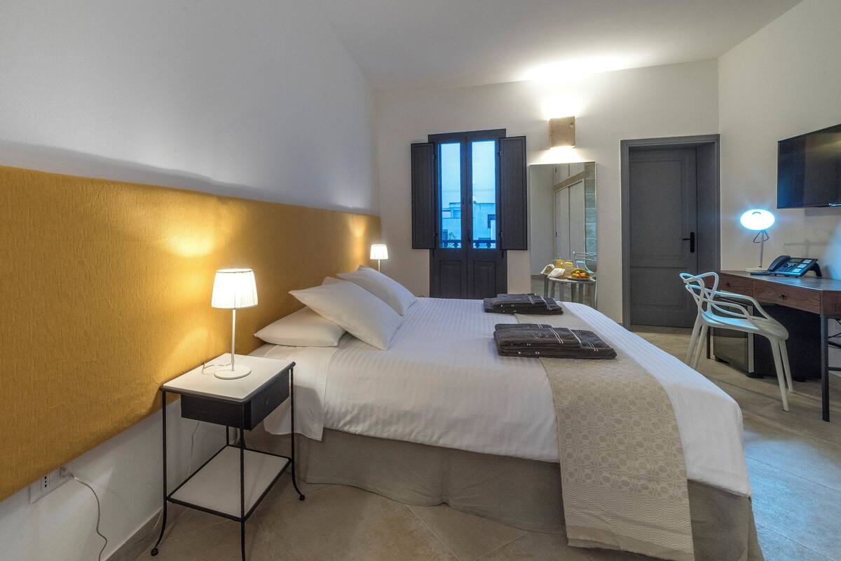 A hotel room with a bed, bedside table, lamp, desk with chair, and a TV.