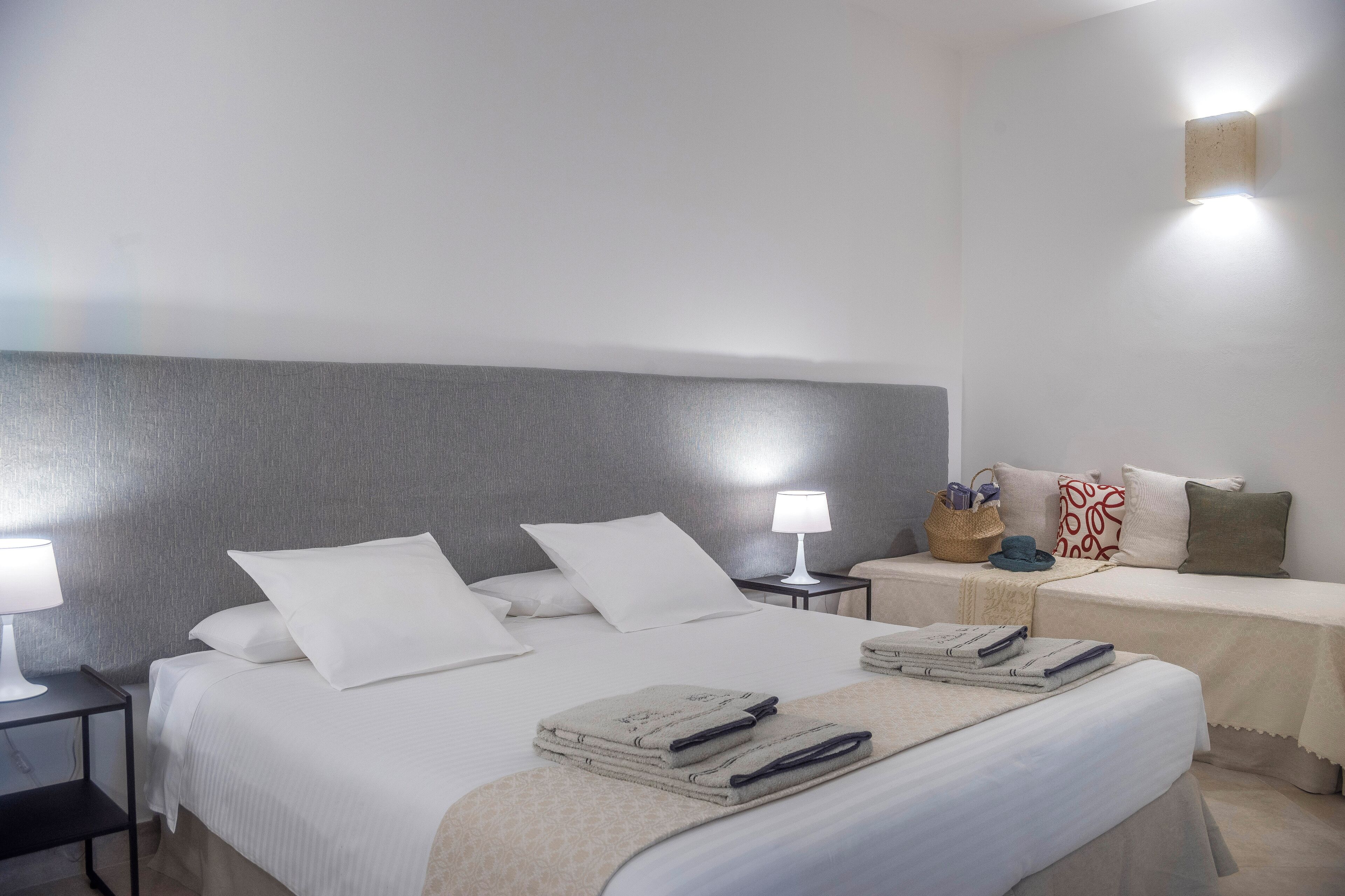 Una cama bien hecha con sábanas blancas, una lámpara de noche y una mesita con lámpara y cesta.