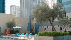 A swimming pool with lounge chairs and umbrellas, surrounded by modern buildings and a tree. - 1BHK. Burj View. Business Bay. Vera II (Dubai)