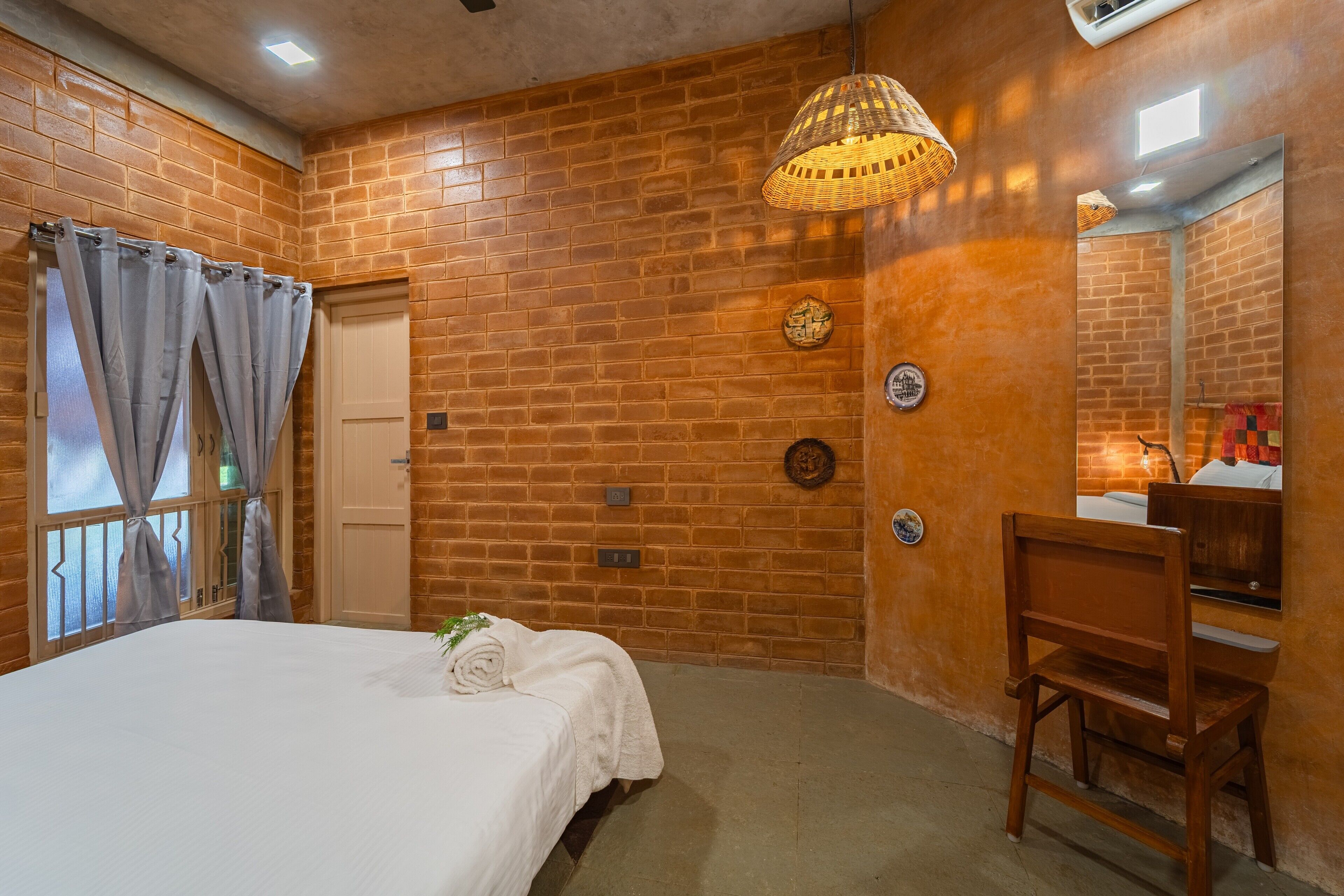 A bedroom with a brick wall, a bed with white linens, a wooden chair, and a mirror.
