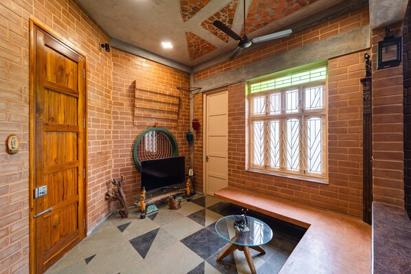 A room with a brick wall, a wooden door, a television, a bench, and a window with bars.