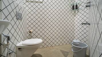 Un baño con un inodoro blanco, un bidé, un pequeño estante con artículos de aseo y un cubo.