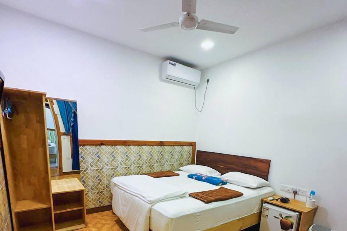 A bedroom with a bed, a ceiling fan, an air conditioning unit, a small refrigerator, and a wooden wardrobe.