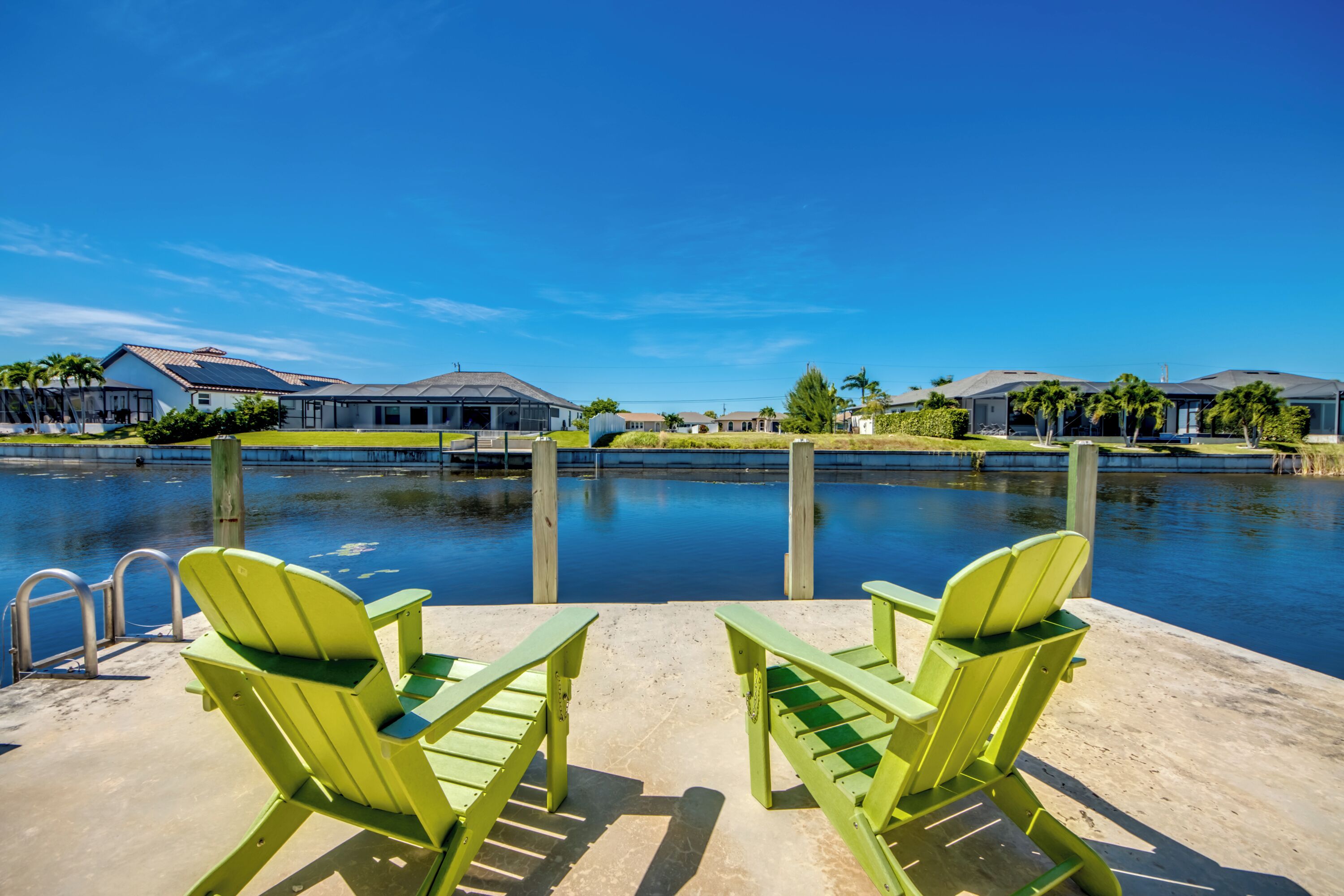 Un espace aménagé au bord de la piscine, avec deux fauteuils Adirondack verts, une terrasse en béton et une vue sur les maisons de l’autre côté de l’eau.