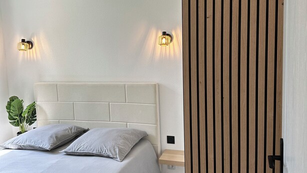 Quarto com cama, duas luminárias na parede, mesa de cabeceira, planta e painel de madeira na parede.