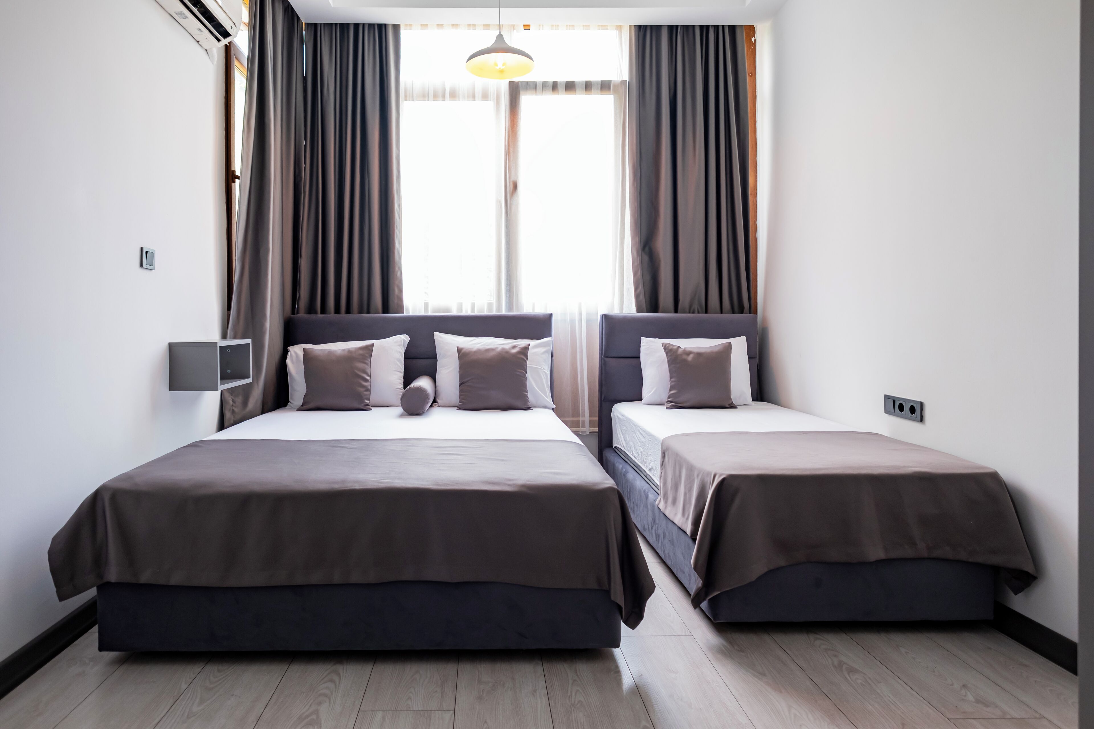 A double room with two beds, a large window with curtains, a wall-mounted air conditioner, and a pendant light.