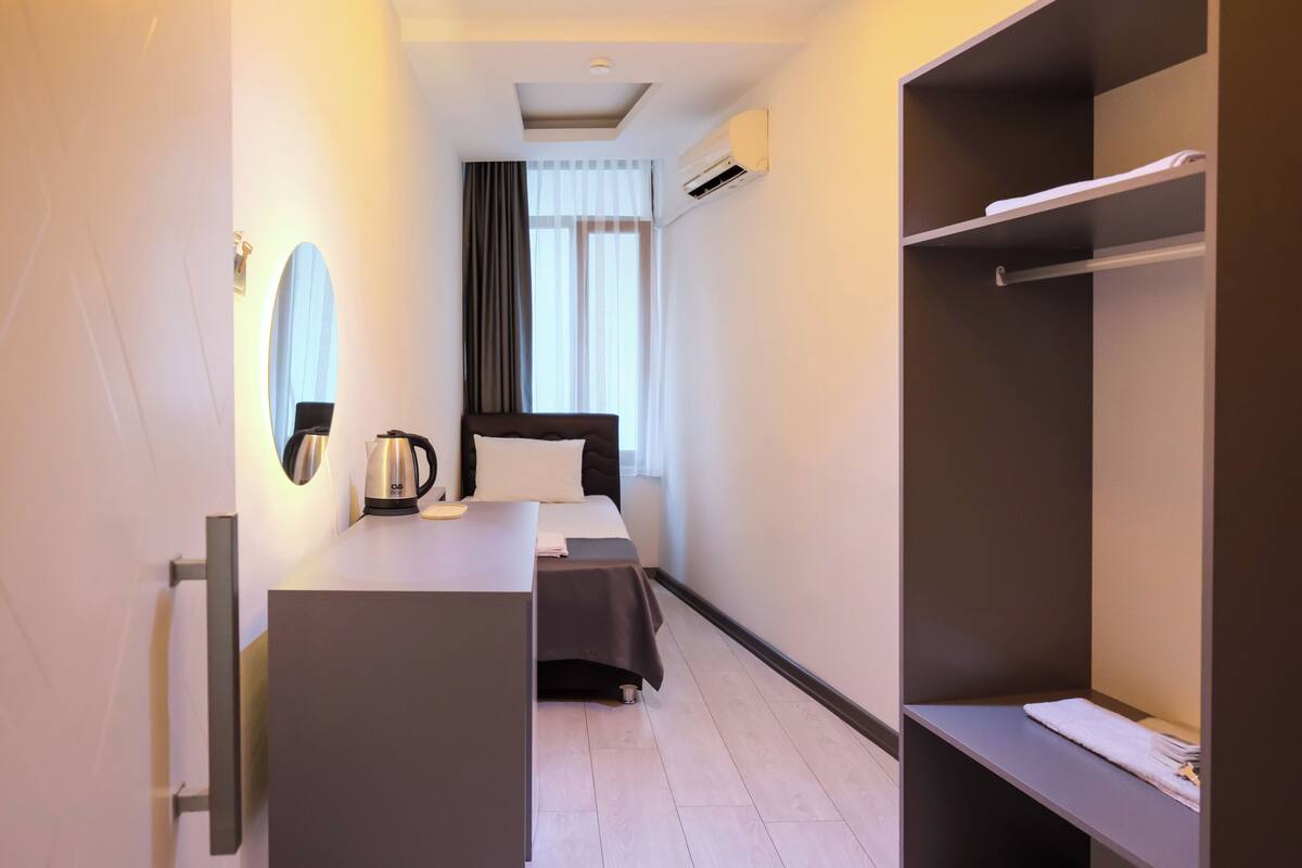 A hotel room with a bed, a desk with a kettle, a wardrobe, and a window with curtains.