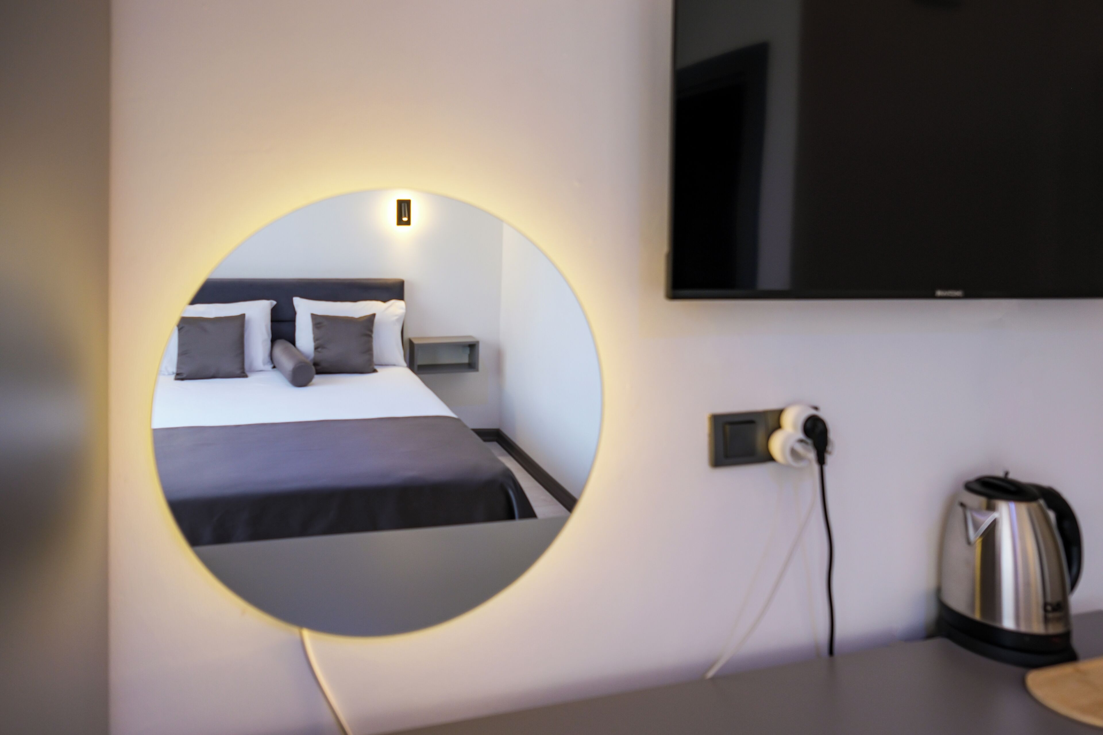 A modern hotel room with a circular mirror, a bed with black bedding, a flat-screen TV mounted on the wall, a kettle, and a power outlet.