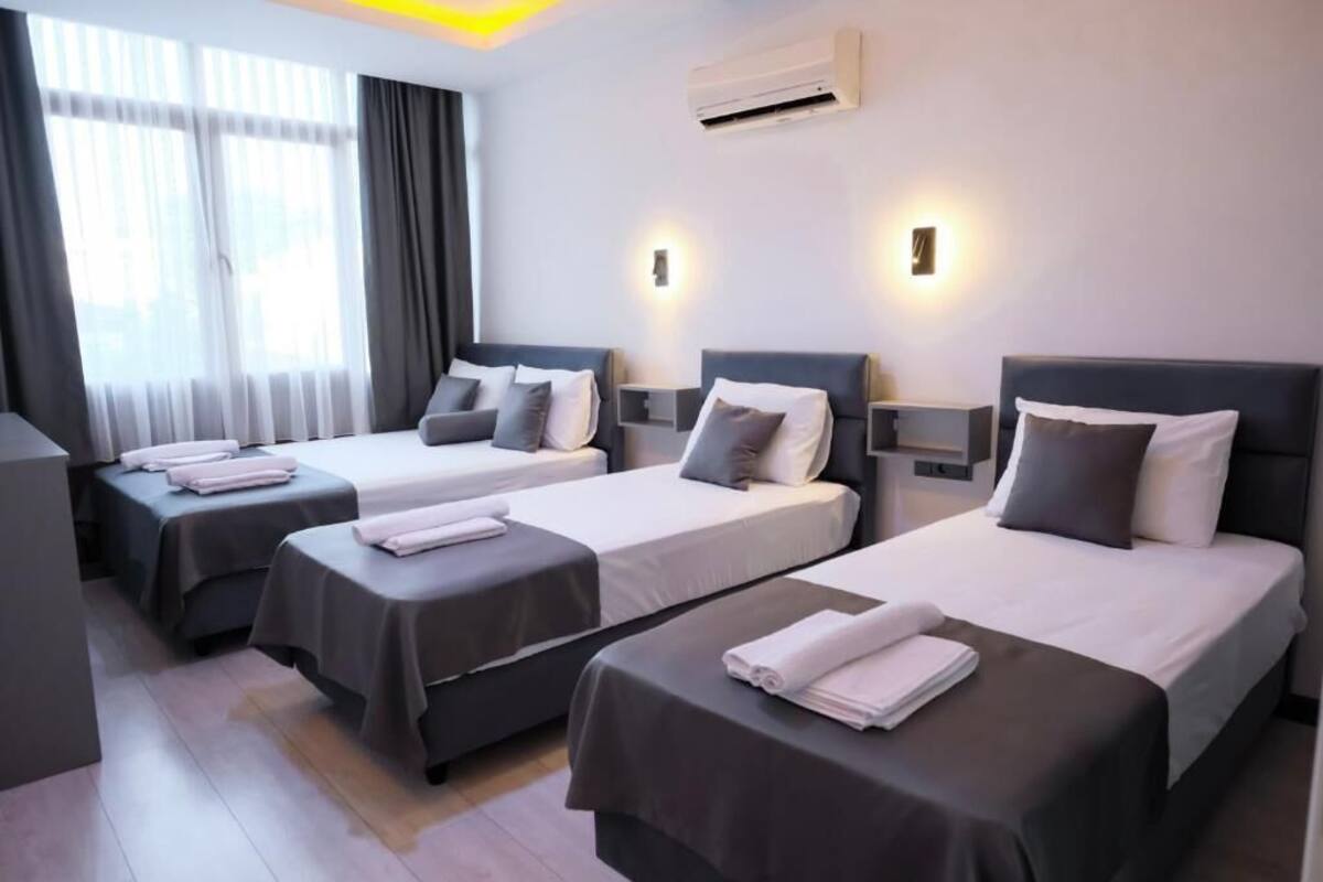 A hotel room with three beds, each with a pillow and a folded towel. There is an air conditioning unit on the wall and a window with curtains.