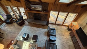 A spacious living room with a stone fireplace, wooden ceiling beams, and large windows offering a view of the outdoors. - 3-bedroom mountain oasis in Black Butte Ranch surrounded by trees (Sisters)