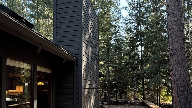 A wooden deck with benches, a building with large windows, and a forested background. - 3-bedroom mountain oasis in Black Butte Ranch surrounded by trees (Sisters)