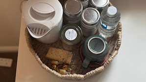 Kurv med hårtørrer, krukker, en kop og lidt snacks på en hvid overflade.