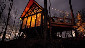 Une cabane en bois dotée de grandes fenêtres et d’une terrasse, entourée d’arbres et illuminée de guirlandes lumineuses.
