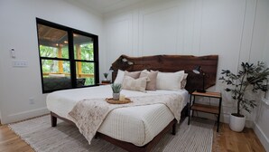Une chambre avec une tête de lit en bois, une grande fenêtre, une table de chevet, une commode, une plante en pot et une lampe de chevet.