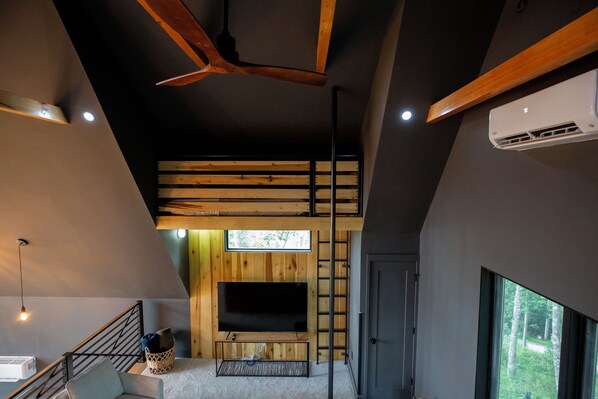 A modern interior with a loft-style ceiling, a built-in TV, and a large window offering a view of greenery. - Luxury Treehouse with Sauna, Hot Tub, and Mountain Views (Vilas)