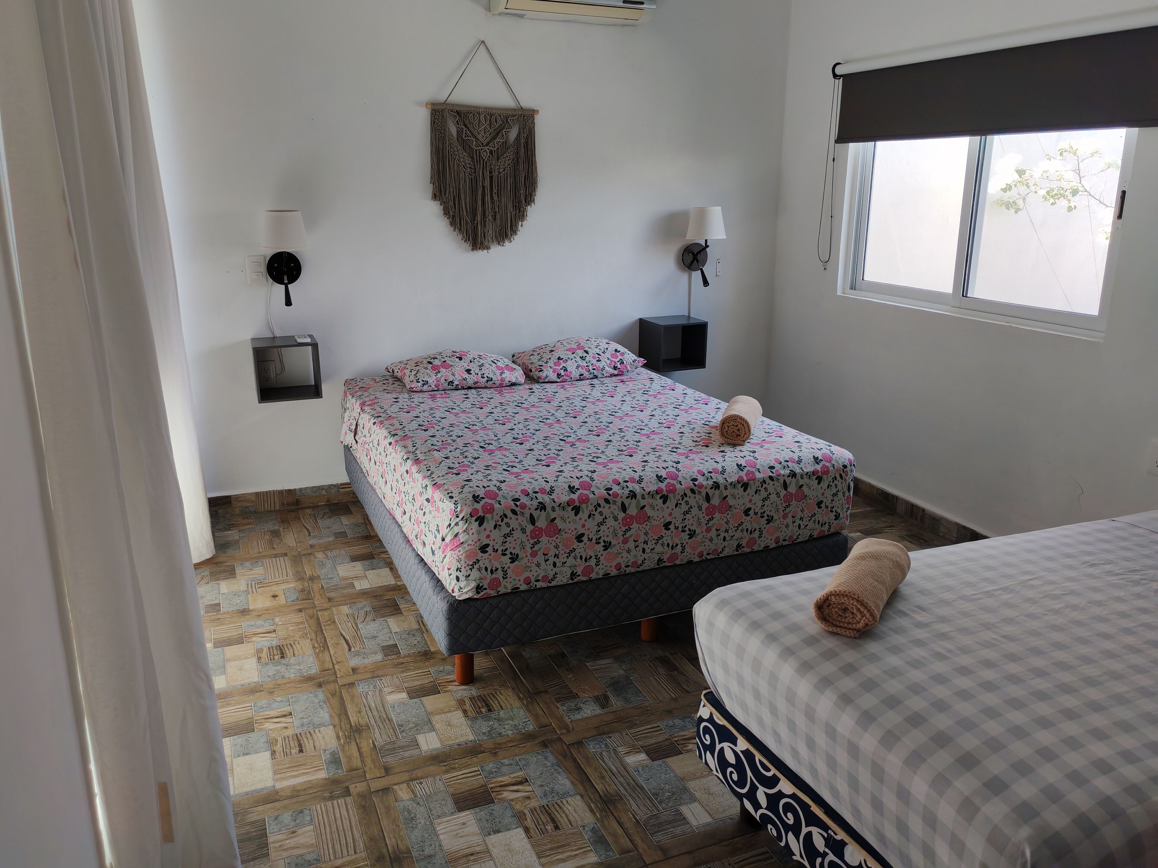 A bedroom with a bed, a smaller bed, a wall-mounted macrame piece, two wall sconces, and a window.