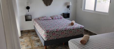 A bedroom with a bed, a smaller bed, a wall-mounted macrame piece, two wall sconces, and a window.