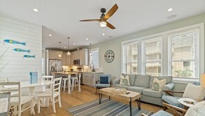 A spacious living room with a sofa, coffee table, and dining area. There is a kitchen in the background with white cabinets and a ceiling fan. - Carolina Skies - Rooftop Deck - Ocean Views (Carolina Beach)