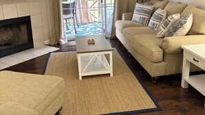 Una sala de estar con chimenea, ventilador de techo, sofá, mesa de centro y vistas a un patio exterior a través de un ventanal.