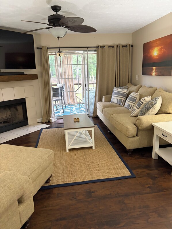 A living room with a fireplace, a ceiling fan, a sofa, a coffee table, and a view through a glass door to an outdoor patio. - Lake Martin Condo in StillWaters (Dadeville)